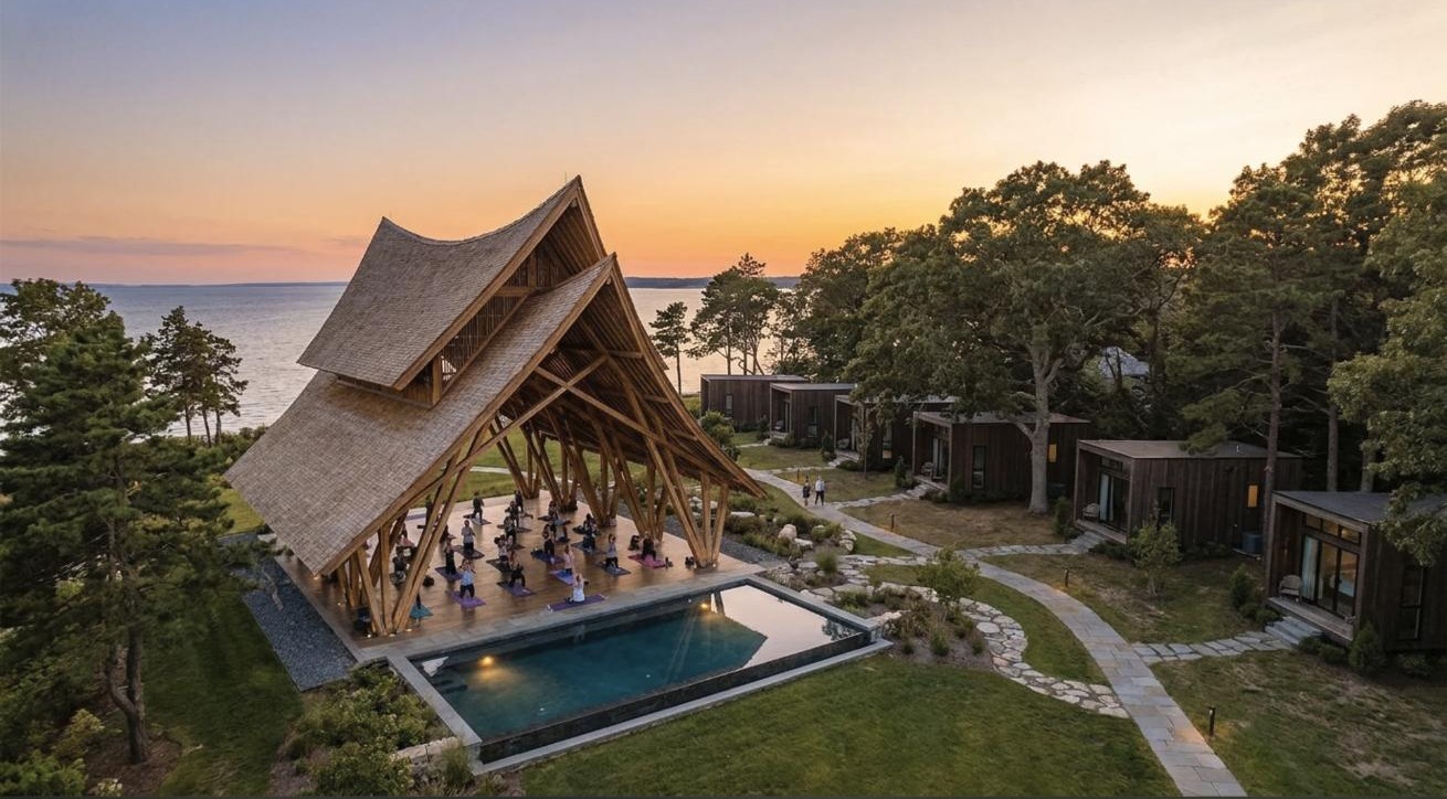 Aerial view of a timber ceremonial pavilion with a yoga class, pool and guest cabins at sunset on the shoreline